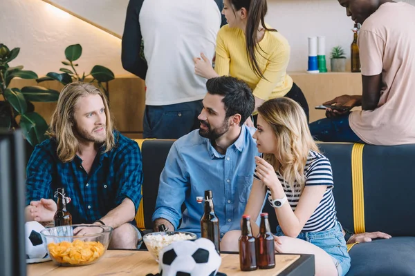 Cropped image of group multicultural football fans sitting on sofa and talking to each other near table with beer, chips and popcorn — Stock Photo