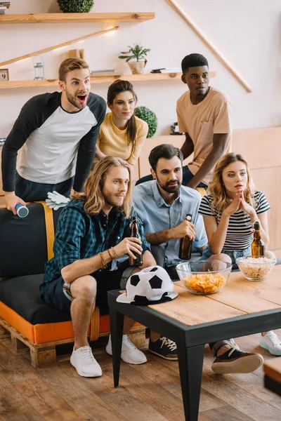 Jovens amigos multiculturais com garrafas de cerveja assistindo jogo de futebol em casa — Fotografia de Stock