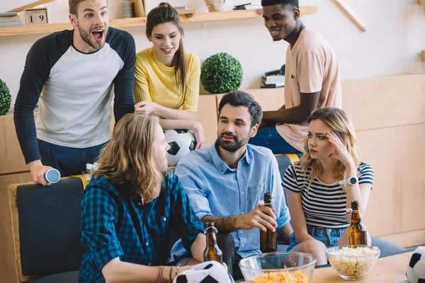 Amigos multiculturales con pelota de fútbol y botellas de cerveza hablando y viendo el partido de fútbol en casa - foto de stock