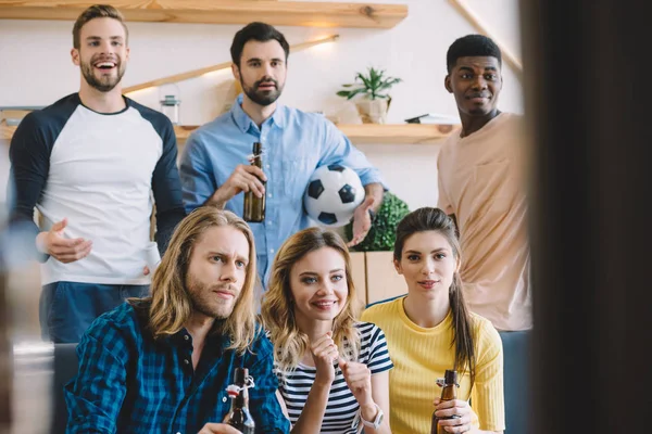 Група мультикультурних друзів з футбольним м'ячем і пляшками пива, які спостерігають футбольний матч вдома — стокове фото