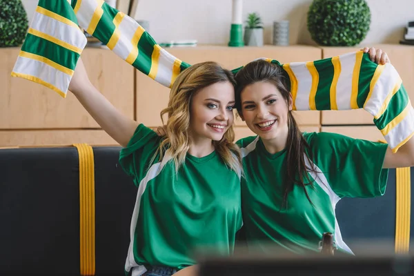 Улыбающиеся женщины-футбольные болельщицы в зеленых футболках и шарфе, празднующие во время просмотра футбольного матча дома — стоковое фото