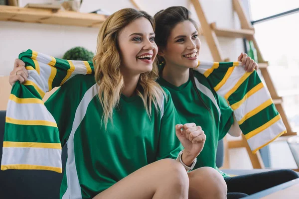 Happy female football fans in green t-shirts and scarf gesturing by hands during watch of soccer match at home — Stock Photo