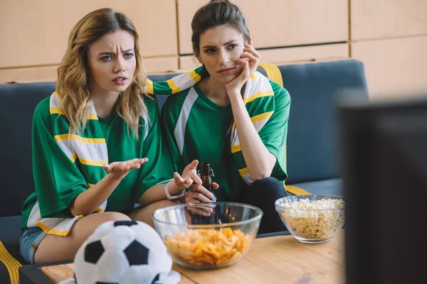 Две расстроенные футбольные фанатки в зеленых футболках и шарфе, жестикулирующие руками во время просмотра футбольного матча дома — стоковое фото