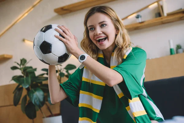 Jovem fã de futebol feminino animado em t-shirt verde e cachecol segurando bola e celebrando durante o relógio de jogo de futebol em casa — Fotografia de Stock