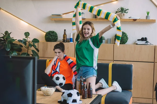 Mujer emocionada en verde ventilador camiseta celebrando la victoria y sosteniendo bufanda sobre la cabeza mientras que su amiga molesta en naranja camiseta sentada cerca en el sofá durante el reloj del partido de fútbol en casa - foto de stock