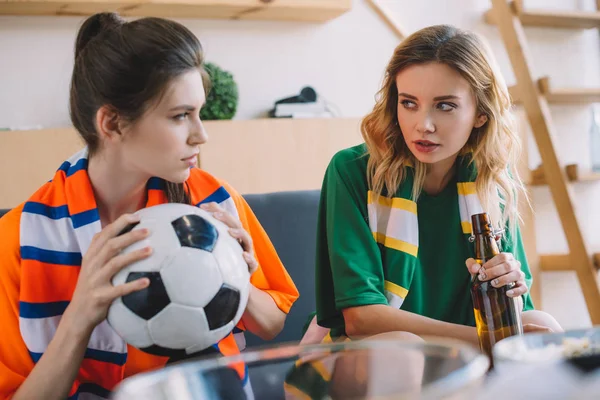 Two female football fans in different fan t-shirts with ball and beer bottle looking at each other at home — Stock Photo