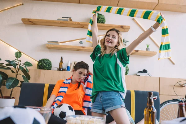 Молодая улыбающаяся женщина в зеленой футболке, празднующая победу и держащая шарф над головой, в то время как ее расстроенная подруга в оранжевой футболке сидит позади во время просмотра футбольного матча дома — стоковое фото