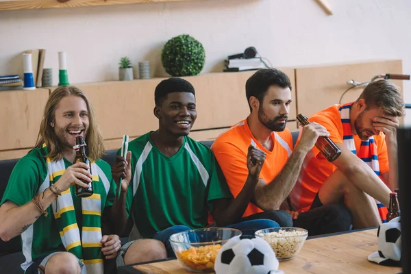 Улыбающиеся футбольные фанаты в зеленых футболках празднуют, в то время как их расстроенные друзья в оранжевых футболках сидят на диване во время просмотра футбольного матча дома — стоковое фото