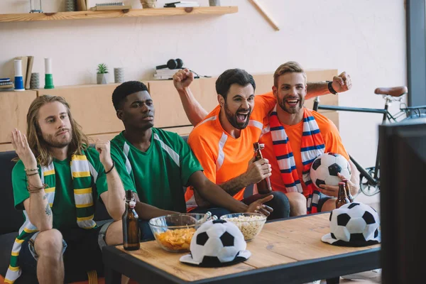 Torcedores de futebol felizes em camisetas laranja comemorando enquanto seus amigos chateados em camisetas verdes sentados perto do sofá durante o relógio de jogo de futebol em casa — Fotografia de Stock