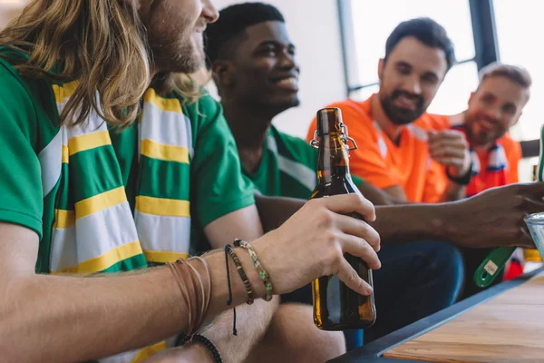 Cortado tiro de homem em verde fã t-shirt e cachecol segurando garrafa de cerveja, enquanto seus amigos masculinos multiculturais assistindo jogo de futebol em casa — Fotografia de Stock
