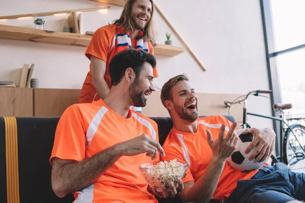 Sonrientes fanáticos del fútbol masculino en camisetas naranjas con pelota y palomitas de maíz viendo el partido de fútbol en casa - foto de stock