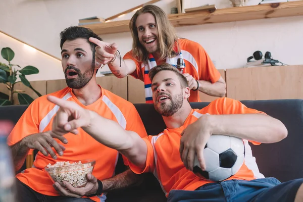 Jóvenes fanáticos del fútbol masculino en camisetas naranjas señalando con los dedos a un amigo sorprendido con palomitas de maíz durante el reloj del partido de fútbol en casa - foto de stock