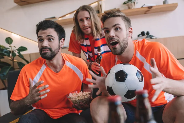 Emocionales jóvenes fanáticos del fútbol masculino en camisetas naranjas con pelota y palomitas de maíz viendo el partido de fútbol en casa - foto de stock