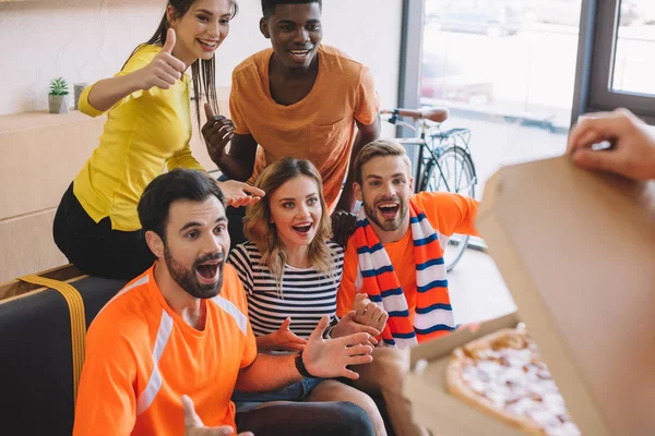 Cropped shot of person holding pizza and excited group of multicultural friends at home — Stock Photo
