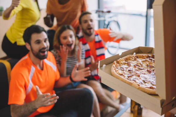 Close up shot of pizza in box and excited group of friend sitting on sofa at home — Stock Photo