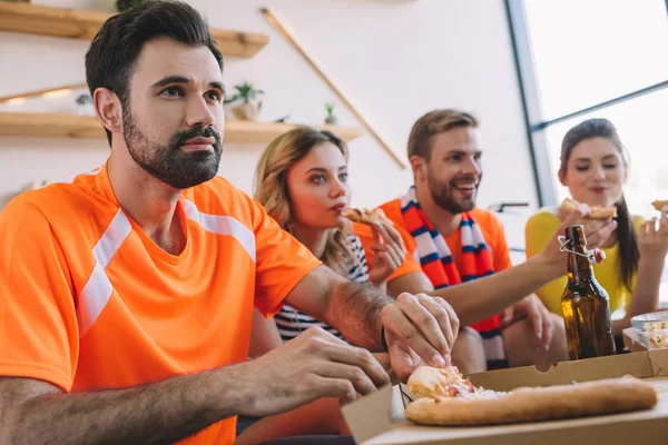 Jeune homme prenant des tranches de pizza de la boîte pendant que ses amis mangent de la pizza et regardent le match de football à la maison — Photo de stock
