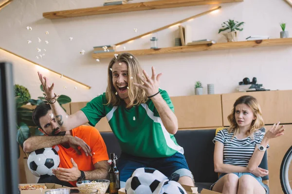 Aufgeregter junger Mann in grünem Fan-T-Shirt, der Popcorn wirft und feiert, während seine aufgebrachten Freunde hinter ihm auf dem Sofa sitzen und gestikulieren, während er das Fußballspiel zu Hause verfolgt — Stockfoto