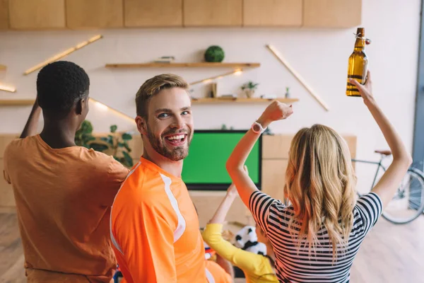 Lächelnder junger Mann blickt in die Kamera, während seine Freunde zu Hause feiern und Fußballspiele auf dem Fernseher verfolgen — Stockfoto