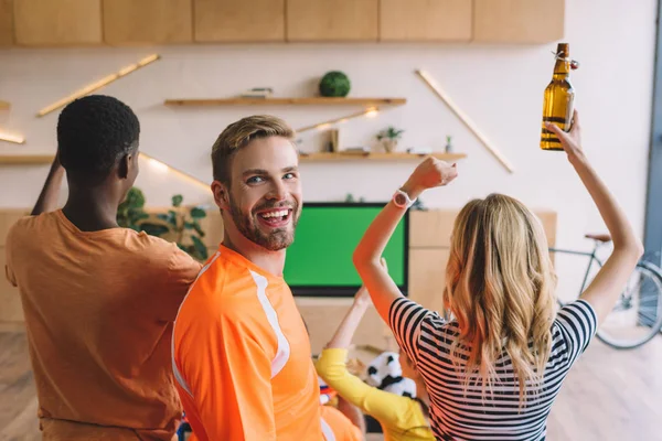 Joven sonriente mirando a la cámara mientras sus amigos celebran y ven el partido de fútbol en la pantalla de televisión en casa - foto de stock