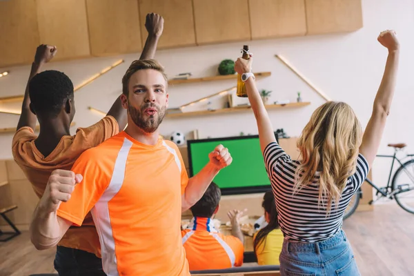 Excité jeune homme faisant des gestes oui et regardant la caméra tandis que ses amis célébrant et regardant match de football sur l'écran de télévision à la maison — Photo de stock