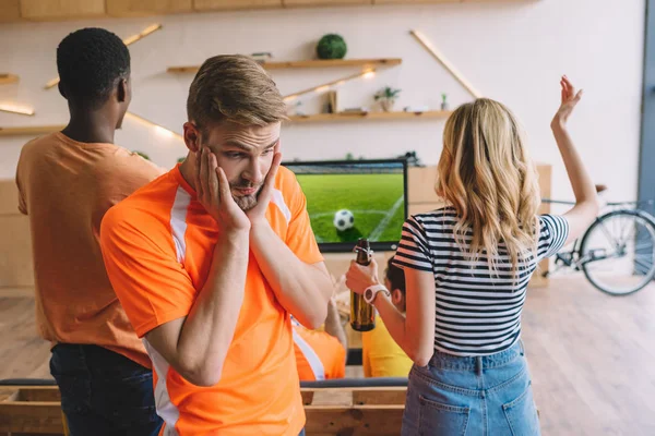 Frustrated young man with hands on cheeks and his friends watching soccer match on tv screen at home — Stock Photo