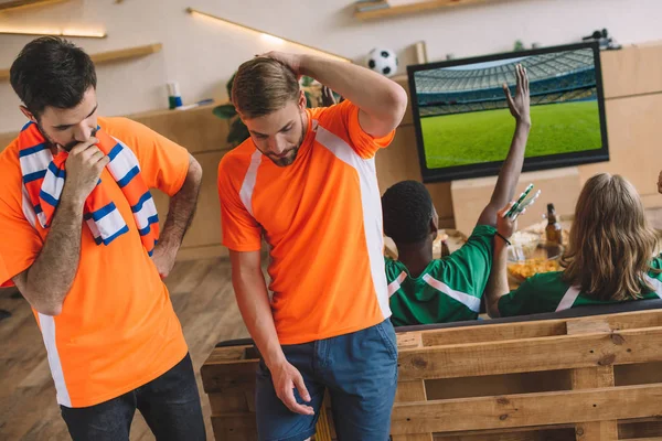 Два расстроенных футбольных фаната в оранжевых футболках и их друзья в зеленых футболках, жестикулирующих и празднующих победу во время просмотра футбольного матча дома — стоковое фото