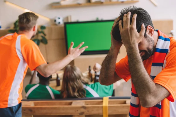 Frustrado joven en naranja camiseta y bufanda cogido de la mano en la cabeza mientras sus amigos viendo el partido de fútbol en la pantalla de televisión en casa - foto de stock