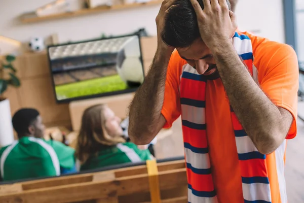 Upset young man in orange t-shirt and scarf holding hands on head while his friends watching soccer match on tv screen at home — Stock Photo