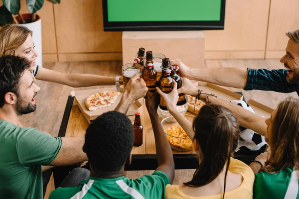 High angle view of football fans celebrating victory and clinking beer bottles and glasses over table with pizza, popcorn and chips at home — Stock Photo