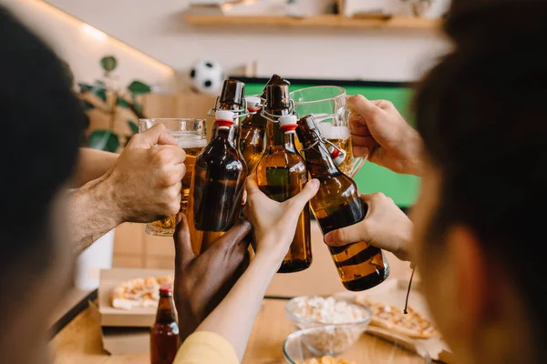 Vista parcial de los aficionados al fútbol celebrando y tintineo botellas de cerveza y vasos sobre la mesa con pizza, palomitas de maíz y patatas fritas en casa - foto de stock