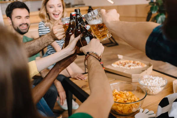 Recortado tiro de los aficionados al fútbol celebrando la victoria y tintineo botellas de cerveza y vasos sobre la mesa con pizza, palomitas de maíz y patatas fritas en casa - foto de stock