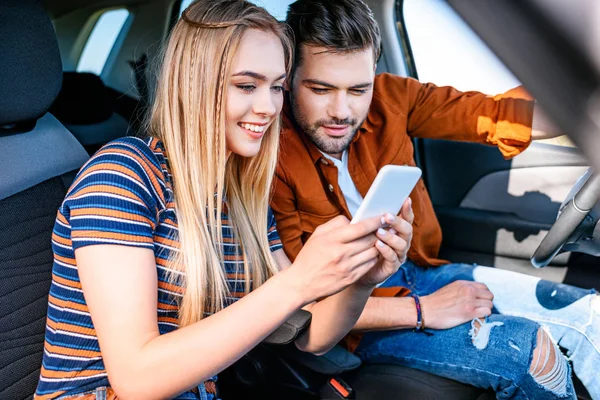 Smiling young couple sitting in car and looking at smartphone screen — Stock Photo