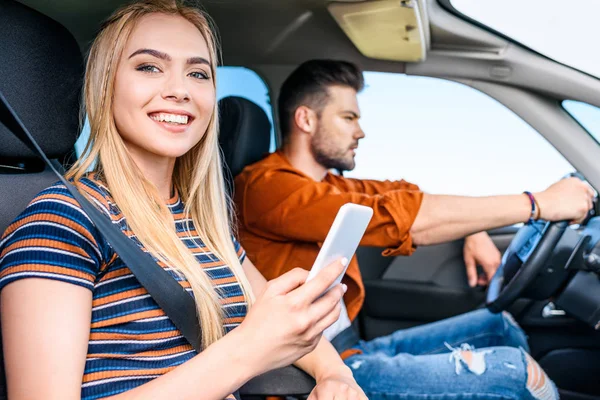 Smiling young woman holding smartphone in hand while her boyfriend driving car — Stock Photo