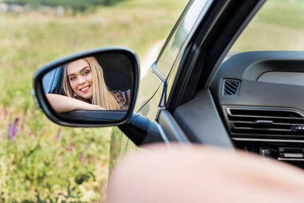 Nahaufnahme einer lächelnden jungen Frau, die im Auto sitzt und in den Seitenspiegel blickt — Stockfoto