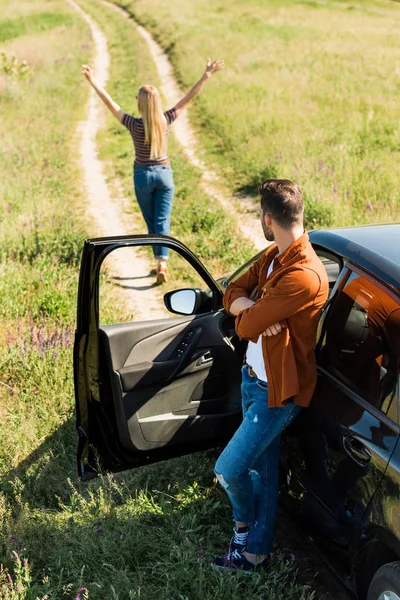 Homme avec les mains croisées debout près de la voiture et regardant petite amie avec des bras larges dans le domaine rural — Photo de stock