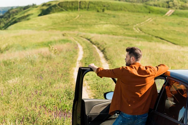 Вид сзади на молодого человека, стоящего рядом с машиной на сельском лугу — стоковое фото