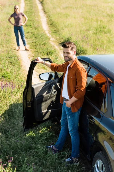 Smiling young man closing door of car and his girlfriend waiting behind on rural meadow — Stock Photo