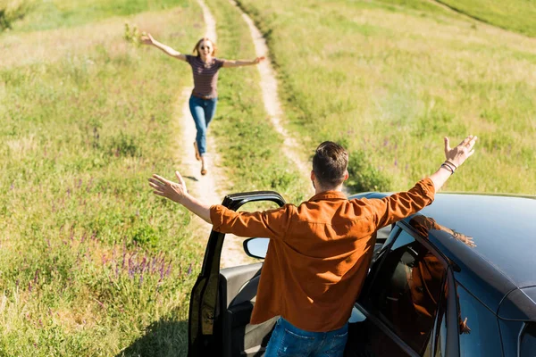 Rear view of man standing with wide arms near car while his girlfriend running to him in field — Stock Photo