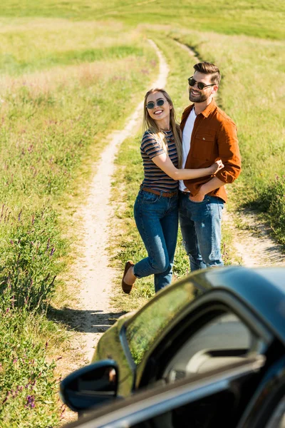 Blick aus der Vogelperspektive auf ein junges stylisches Paar mit Sonnenbrille, das sich in der Nähe eines Autos auf einer ländlichen Wiese umarmt — Stockfoto