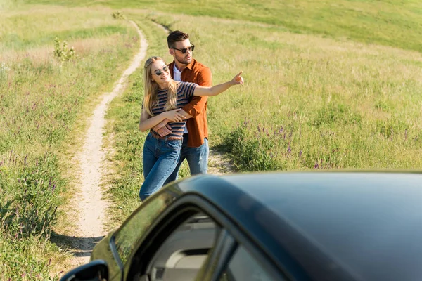 Homem abraçando namorada elegante em óculos de sol apontando por dedo no campo rural perto do carro — Fotografia de Stock