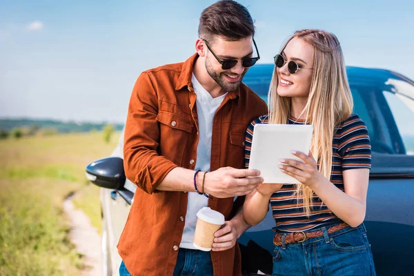 Усміхнена пара в сонцезахисних окулярах стоїть з чашкою кави і цифровим планшетом біля машини — стокове фото
