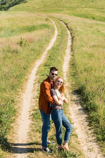 Vista ad alto angolo di felice coppia elegante in occhiali da sole in piedi sul prato rurale — Foto stock