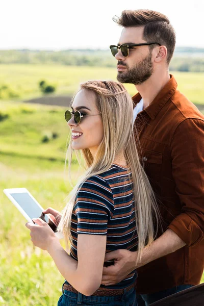 Pareja joven y elegante en gafas de sol de pie con tableta digital - foto de stock