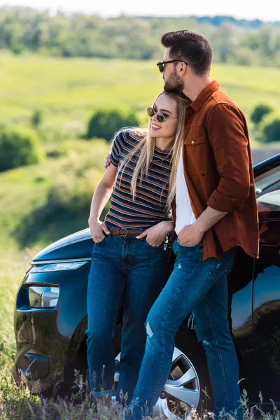 Vista frontal do jovem casal elegante em óculos de sol de pé perto do carro no prado rural — Fotografia de Stock