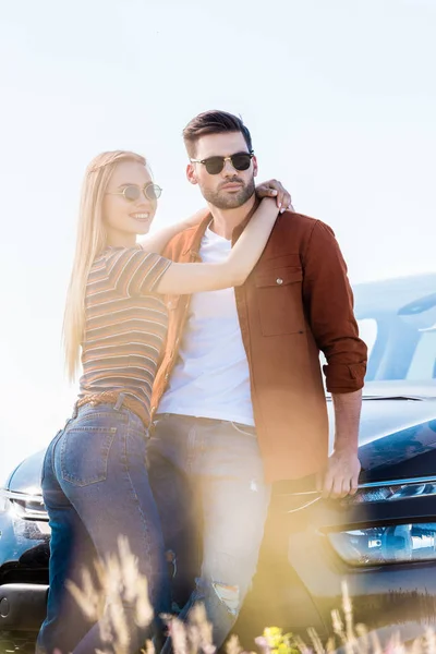 Attractive young woman embracing boyfriend in sunglasses near car — Stock Photo