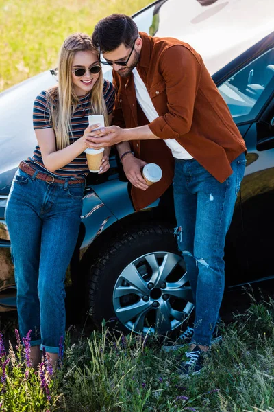 Vista de ángulo alto de la pareja elegante con tazas de café mirando la pantalla del teléfono inteligente cerca del coche - foto de stock