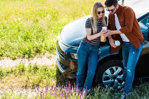 Elevated view of stylish couple with coffee cups looking at smartphone screen near car — Stock Photo