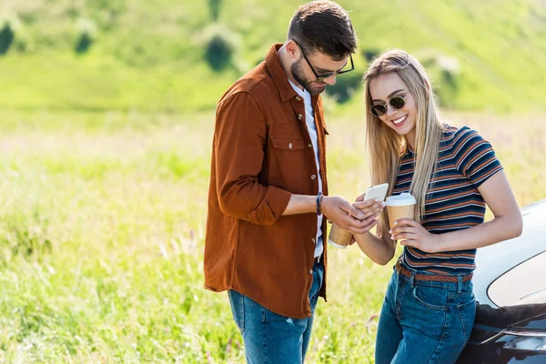 Stylish couple in sunglasses with coffee cups looking at smartphone screen near car — Stock Photo