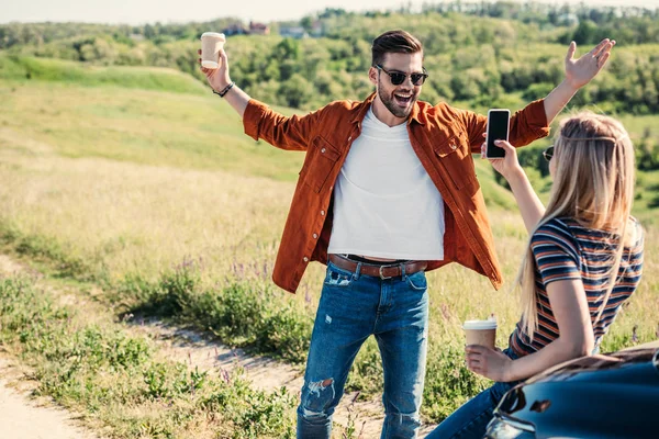 Rear view of woman taking picture of boyfriend with wide arms holding coffee cup near on rural meadow — Stock Photo
