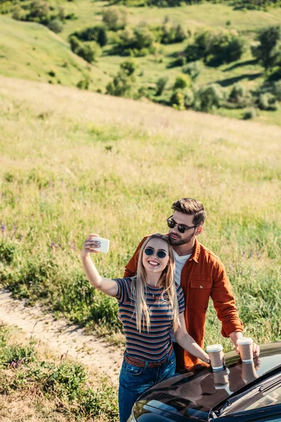 Vista ad alto angolo di coppia elegante in occhiali da sole con tazze di caffè prendere selfie su smartphone vicino auto sul prato rurale — Foto stock